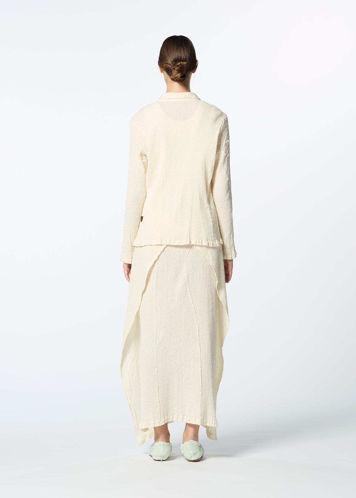 A person stands facing away, wearing HaaT's KYO CHIJIMI EMB cream long-sleeved jacket with textured fabric and a matching uneven-hem skirt. Their hair is tied back, light shoes on, set against a plain background. view 9 of 9