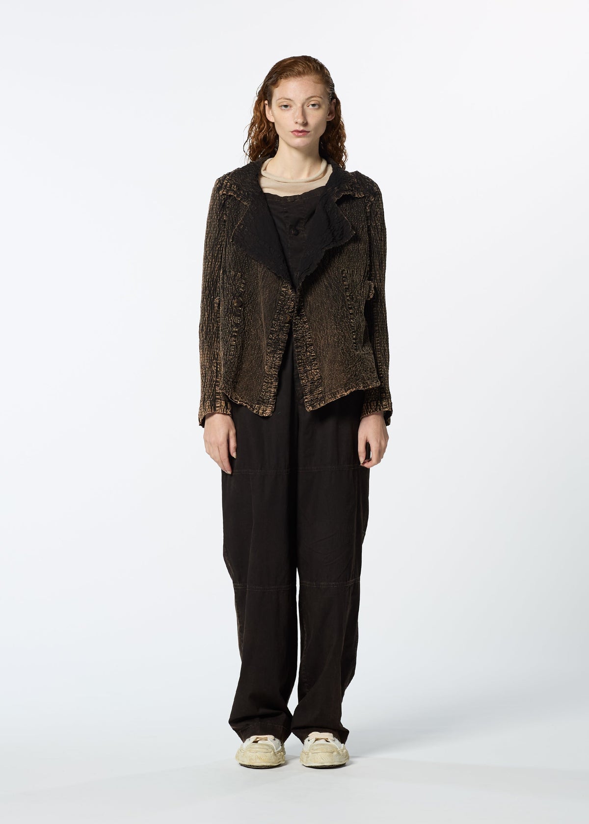 A person with long curly hair faces forward, wearing HaaT's TENMOKU COTTON jacket over a beige top, loose black pants, and white sneakers. The textured garment features subtle three-dimensional shading against a plain white background. view 6 of 9