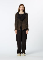 A person with long curly hair faces forward, wearing HaaT's TENMOKU COTTON jacket over a beige top, loose black pants, and white sneakers. The textured garment features subtle three-dimensional shading against a plain white background.