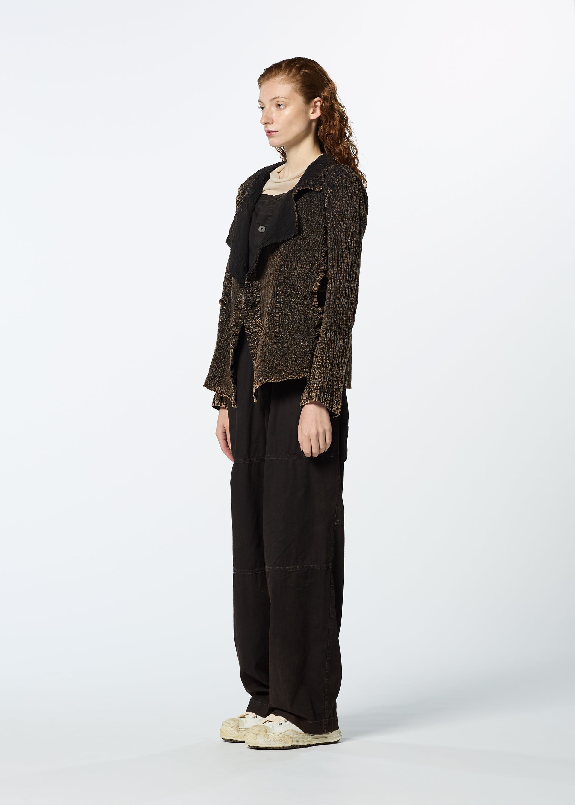 Zoomed view A person with long, wavy red hair stands sideways against a white background, wearing the brown TENMOKU COTTON jacket by HaaT over a black top, loose black pants, and white sneakers.