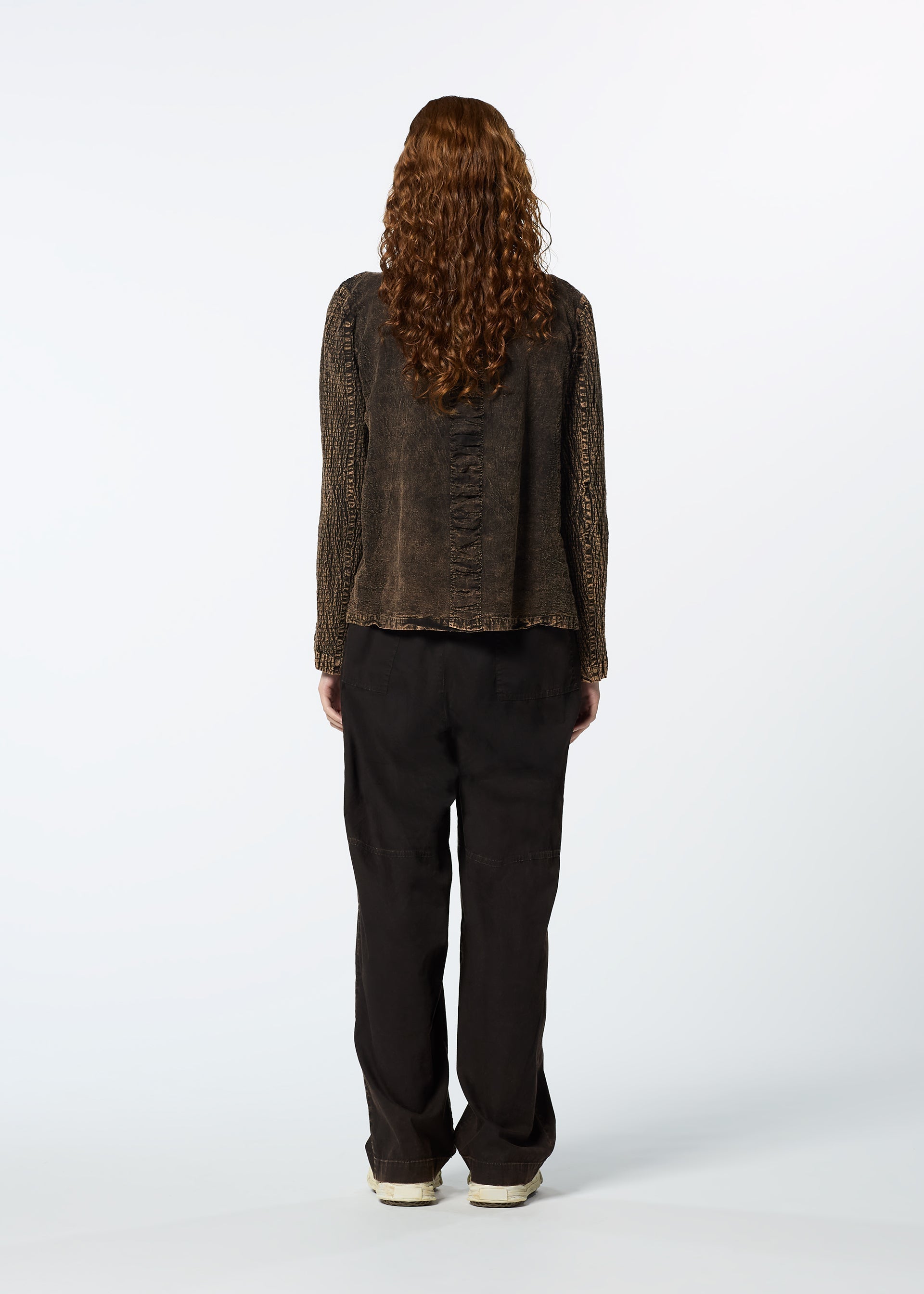 Zoomed view A person with long curly hair, dressed in HaaT’s TENMOKU COTTON top and loose black pants, stands facing away from the camera against a plain white background.