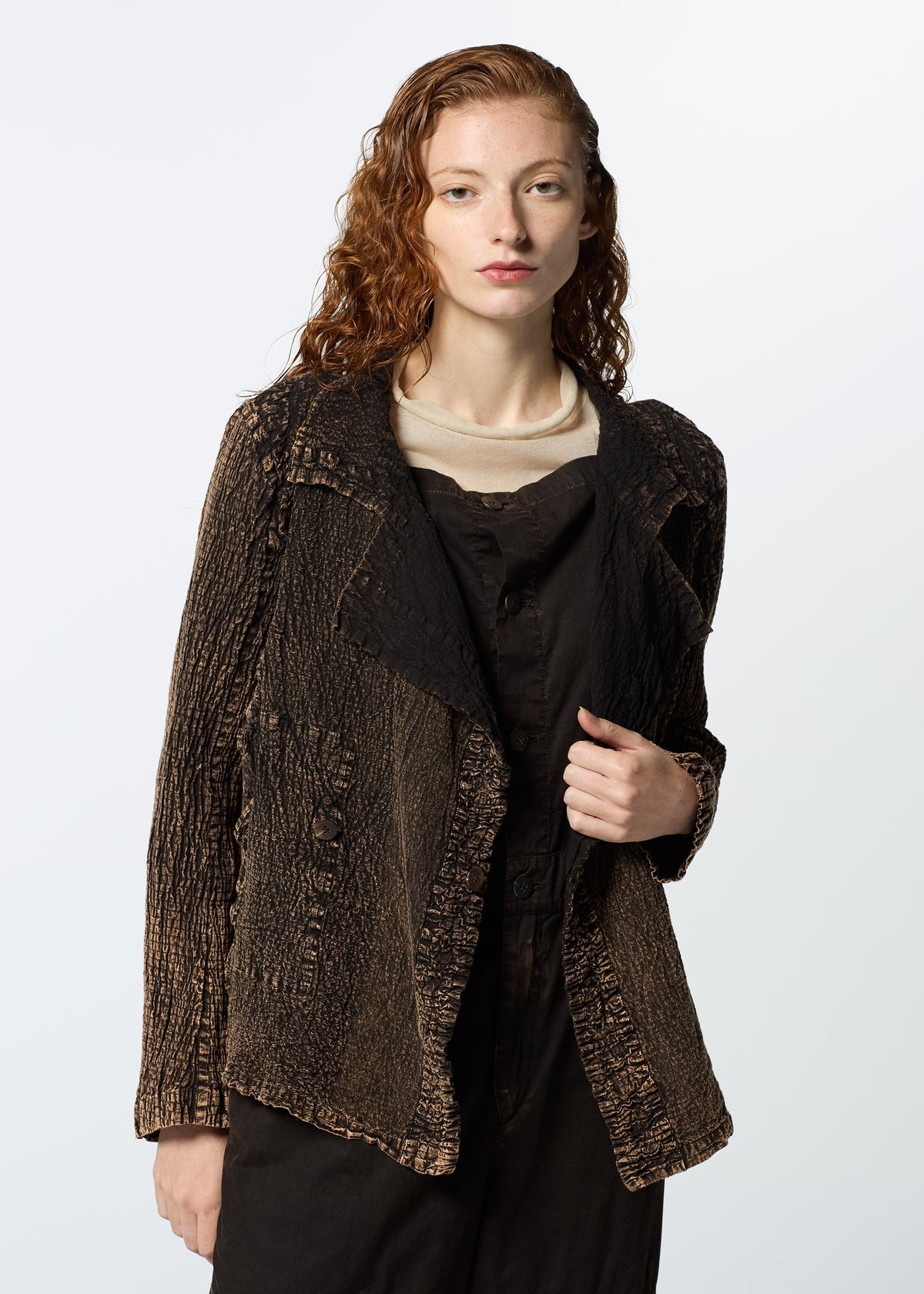 A person with long, wavy reddish hair wears HaaT’s TENMOKU COTTON jacket—a garment-dyed, textured brown piece—over a light top and dark overalls, standing against a plain background and gazing neutrally at the camera. view 9 of 9