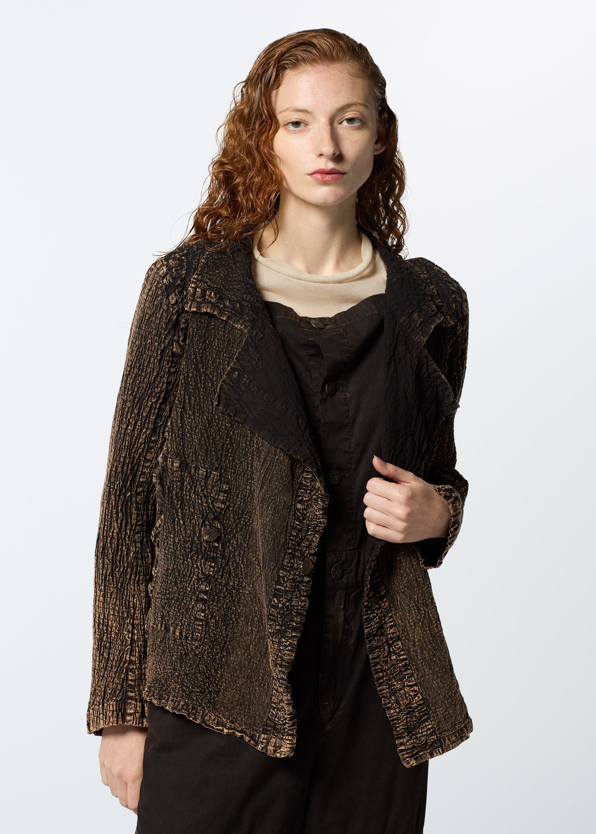 Zoomed view A person with long, wavy reddish hair wears HaaT’s TENMOKU COTTON jacket—a garment-dyed, textured brown piece—over a light top and dark overalls, standing against a plain background and gazing neutrally at the camera.