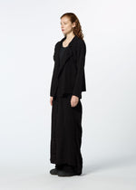 A person with long reddish hair stands in profile against a plain white background wearing HaaT's TENMOKU COTTON garment-dyed, textured black blazer, a black top, ankle-length black skirt, and black shoes.