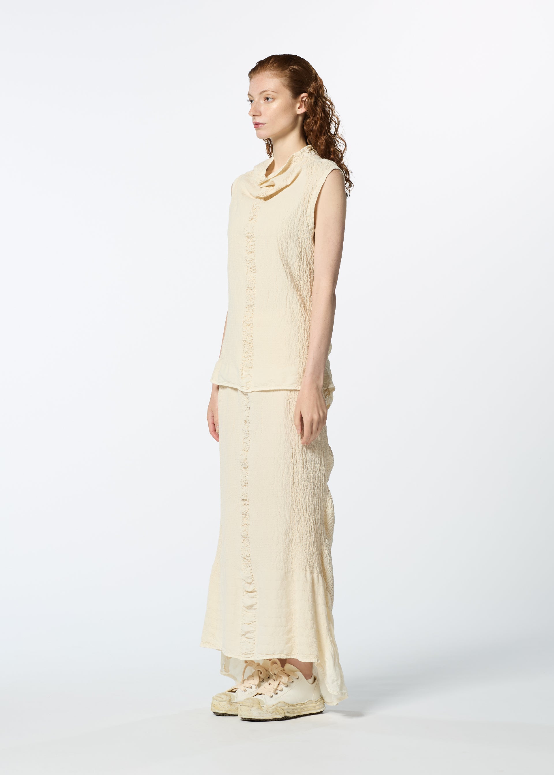 Zoomed view A woman with long, wavy hair poses in profile wearing HaaT's TENMOKU COTTON sleeveless cream garment-dyed top and matching long skirt with cream sneakers, set against a plain white background.