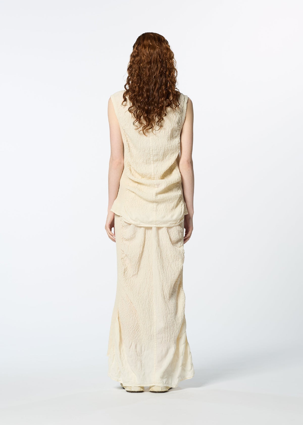 A woman with curly, reddish-brown hair faces away, wearing HaaT’s TENMOKU COTTON textured sleeveless blouse and matching long skirt against a plain white background. view 8 of 8