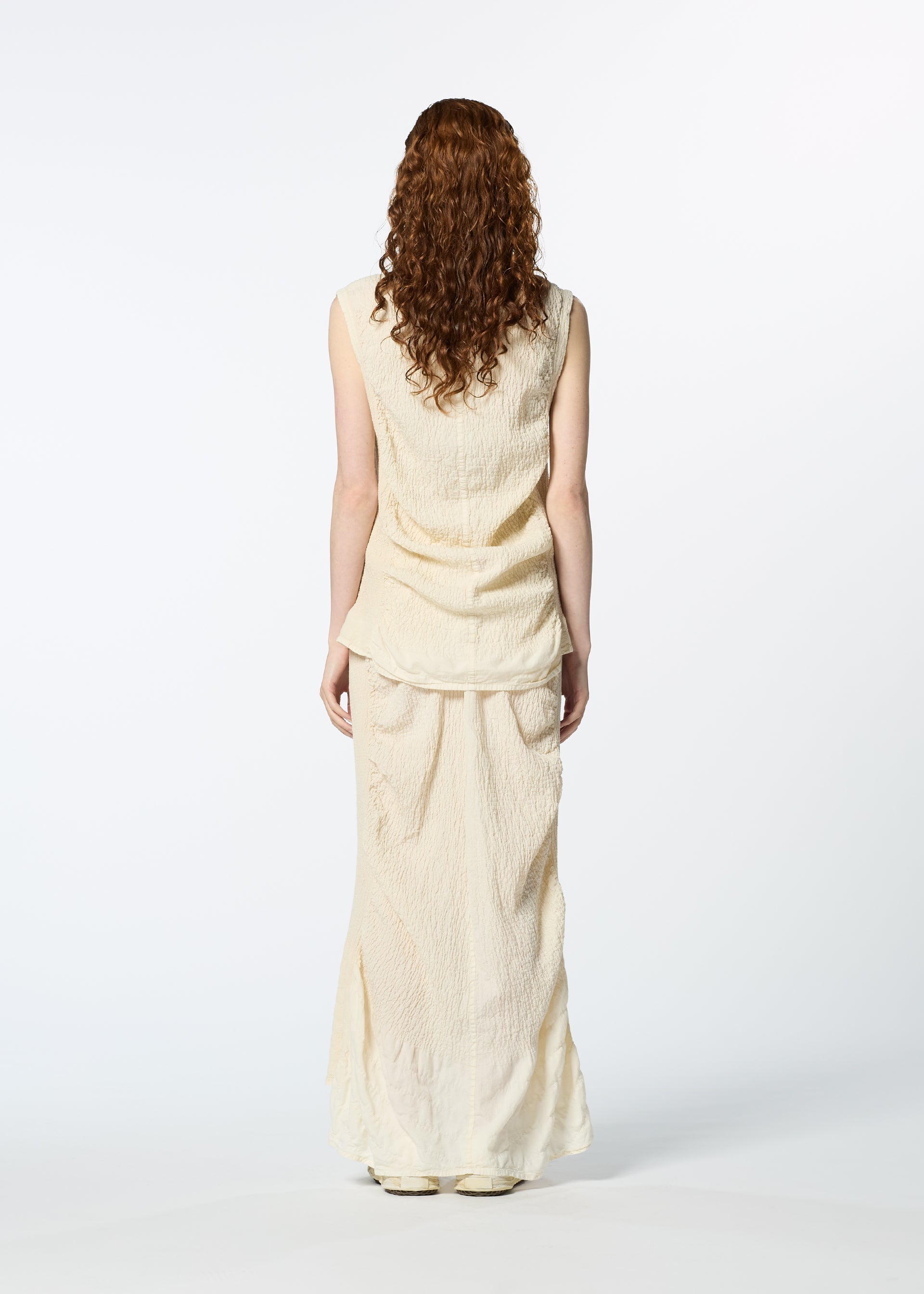 Zoomed view A woman with curly, reddish-brown hair faces away, wearing HaaT’s TENMOKU COTTON textured sleeveless blouse and matching long skirt against a plain white background.