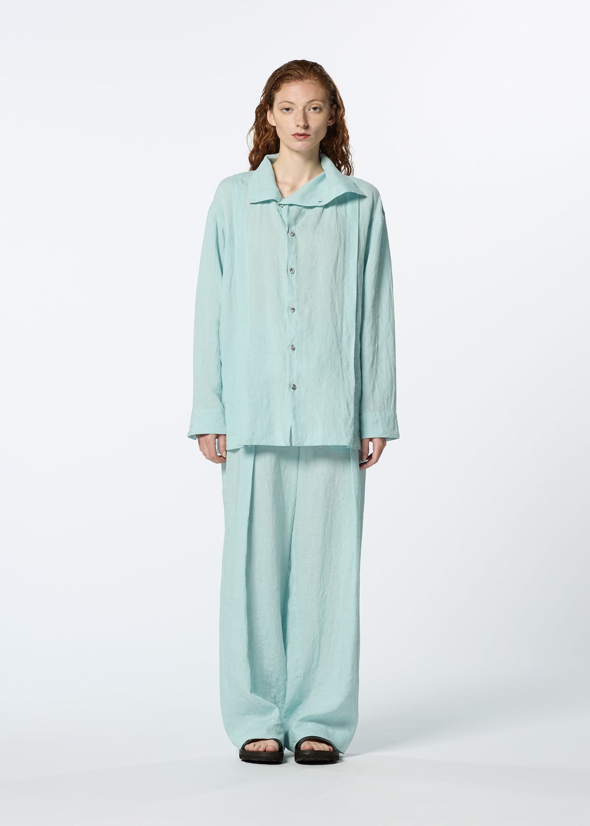 A person with wavy, shoulder-length hair stands facing forward in a loose-fitting, light blue JIKI French linen button-up shirt and matching breathable pants by HaaT, paired with black sandals. The background is plain white. view 6 of 8
