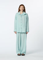 A person with wavy, shoulder-length hair stands facing forward in a loose-fitting, light blue JIKI French linen button-up shirt and matching breathable pants by HaaT, paired with black sandals. The background is plain white.