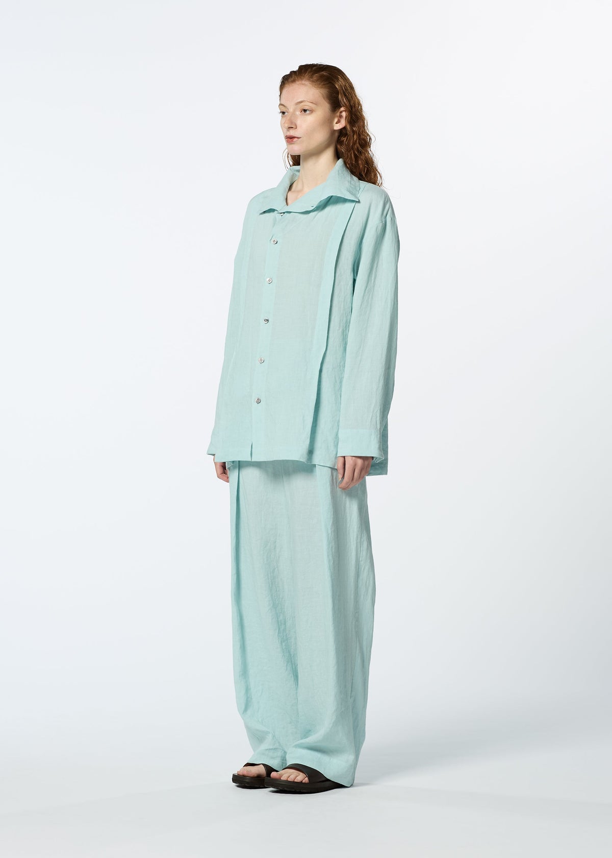 A person stands against a plain background wearing the JIKI light blue French linen button-up shirt and matching breathable pants from HaaT, paired with black sandals for a relaxed, casual look. view 7 of 8