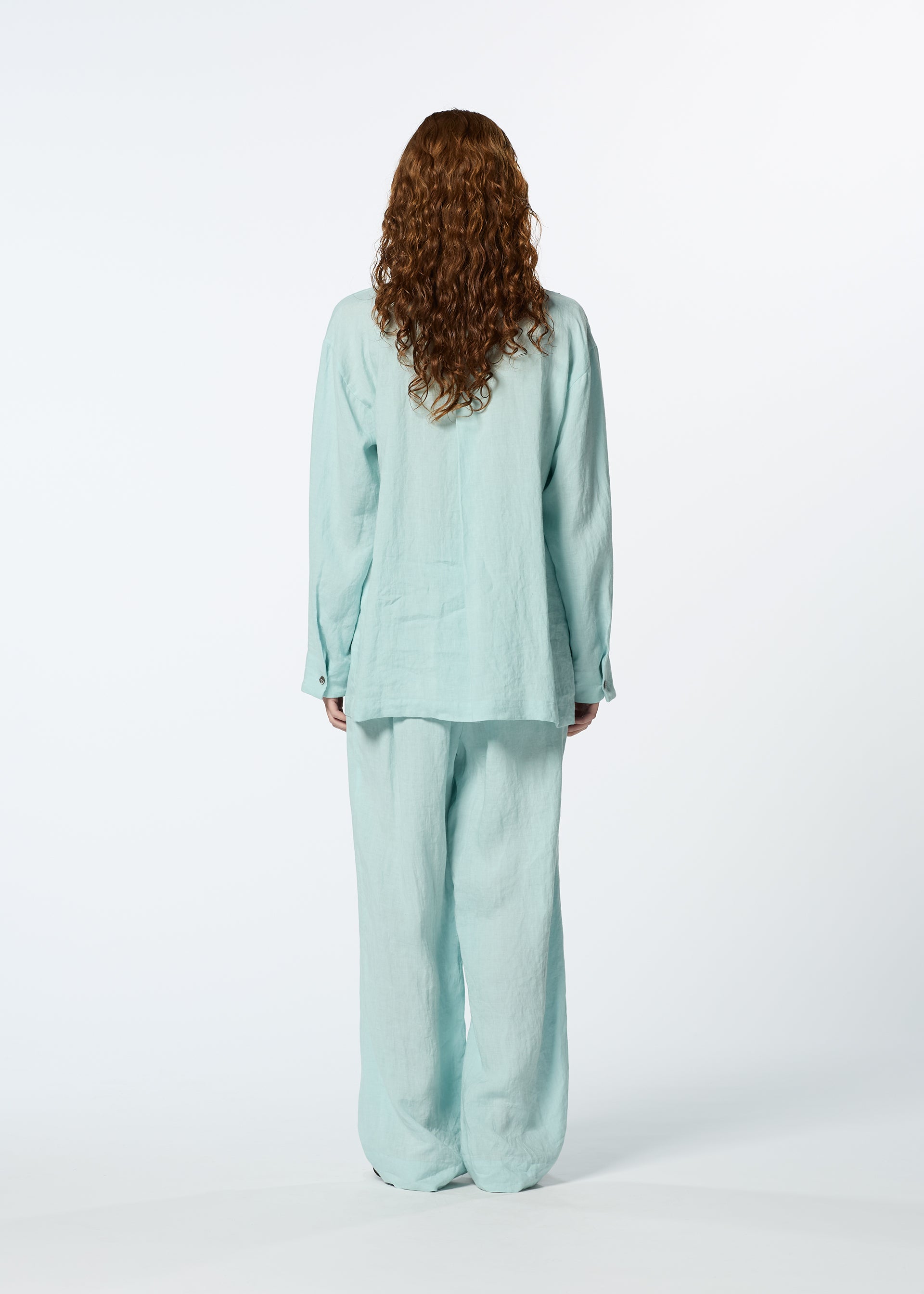 Zoomed view A person with long curly hair, seen from behind, wears the elegant JIKI light blue French linen shirt and breathable pants by HaaT. The loose-fitting silhouette stands out against a plain white background.