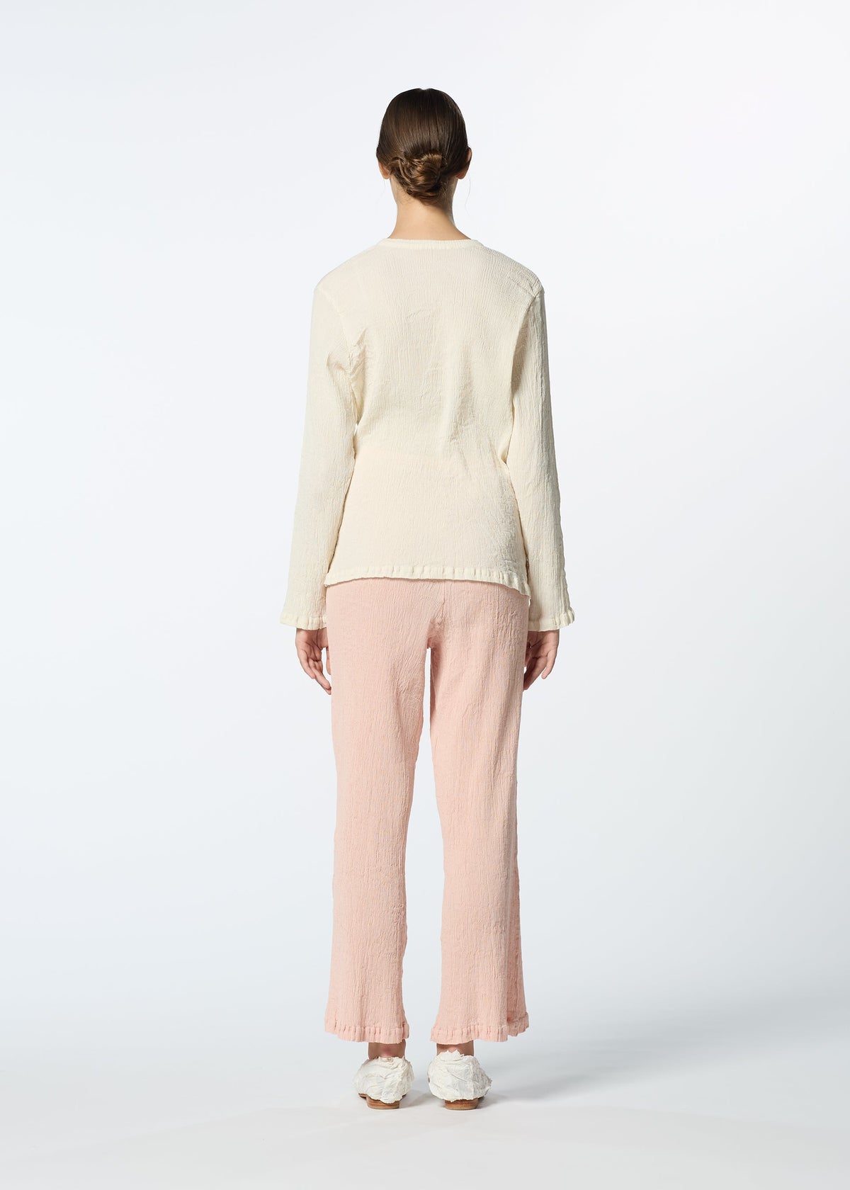 A person stands with their back to the camera in a cream long-sleeve top, HaaT KYO CHIJIMI FEBRUARY ultra-fine cotton pants, and white slippers, hair in a low bun against a plain white background. view 8 of 8