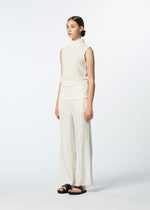 A woman stands against a white background wearing HaaT's SHIBARI BOTTOM wide-leg pleated cream pants, a sleeveless high-neck cream top of hand-folded fabric, and black sandals. Her hair is tied back in a neat bun.