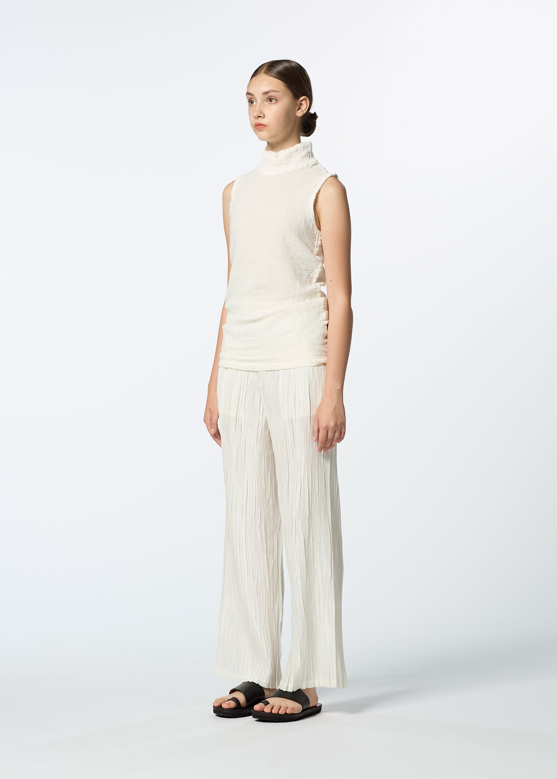 Zoomed view A woman stands against a white background wearing HaaT's SHIBARI BOTTOM wide-leg pleated cream pants, a sleeveless high-neck cream top of hand-folded fabric, and black sandals. Her hair is tied back in a neat bun.