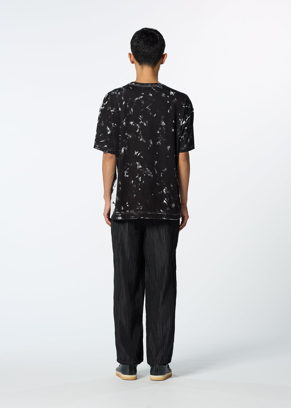 A person faces away from the camera against a white background, wearing HaaT's SHIBARI BOTTOM for a sleek, unisex look with glossy pants styled alongside a black and white tie-dye T-shirt and gray shoes. view 7 of 8