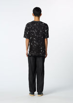 A person faces away from the camera against a white background, wearing HaaT's SHIBARI BOTTOM for a sleek, unisex look with glossy pants styled alongside a black and white tie-dye T-shirt and gray shoes.