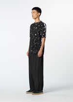 A person stands against a plain white background, wearing a loose black and white patterned T-shirt, HaaT’s SHIBARI BOTTOM wide glossy pants, and black shoes. This unisex look features the individual facing slightly right and looking ahead.