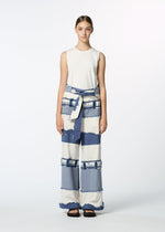 A young woman stands facing forward, wearing HaaT's KASANEGAKE sleeveless white top and jacquard woven blue-and-white striped pants with a matching fabric belt. Her hair is pulled back, she wears black sandals, and poses against a plain background.