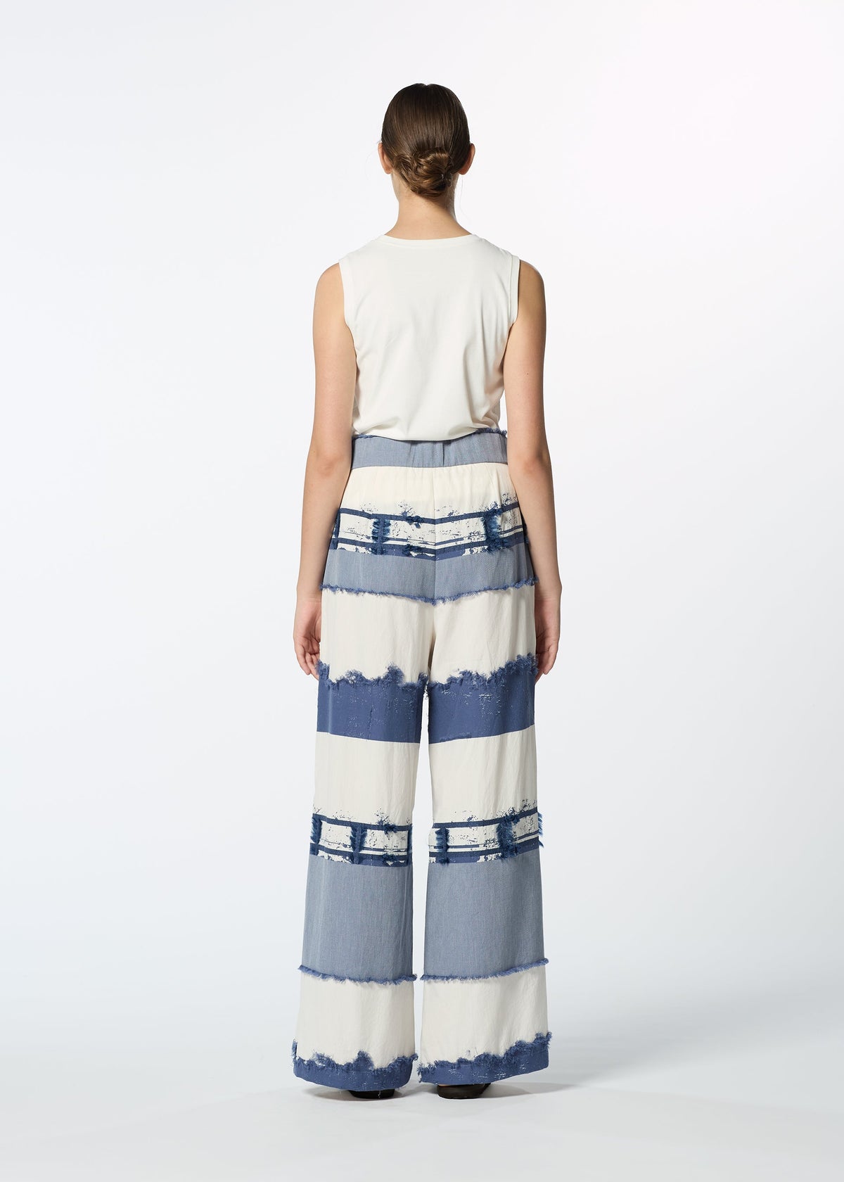 A person stands facing away, wearing HaaT's KASANEGAKE sleeveless white top and jacquard woven wide-leg pants with blue and white stripes, frayed edges, and varied texture, set against a plain white background. view 7 of 7