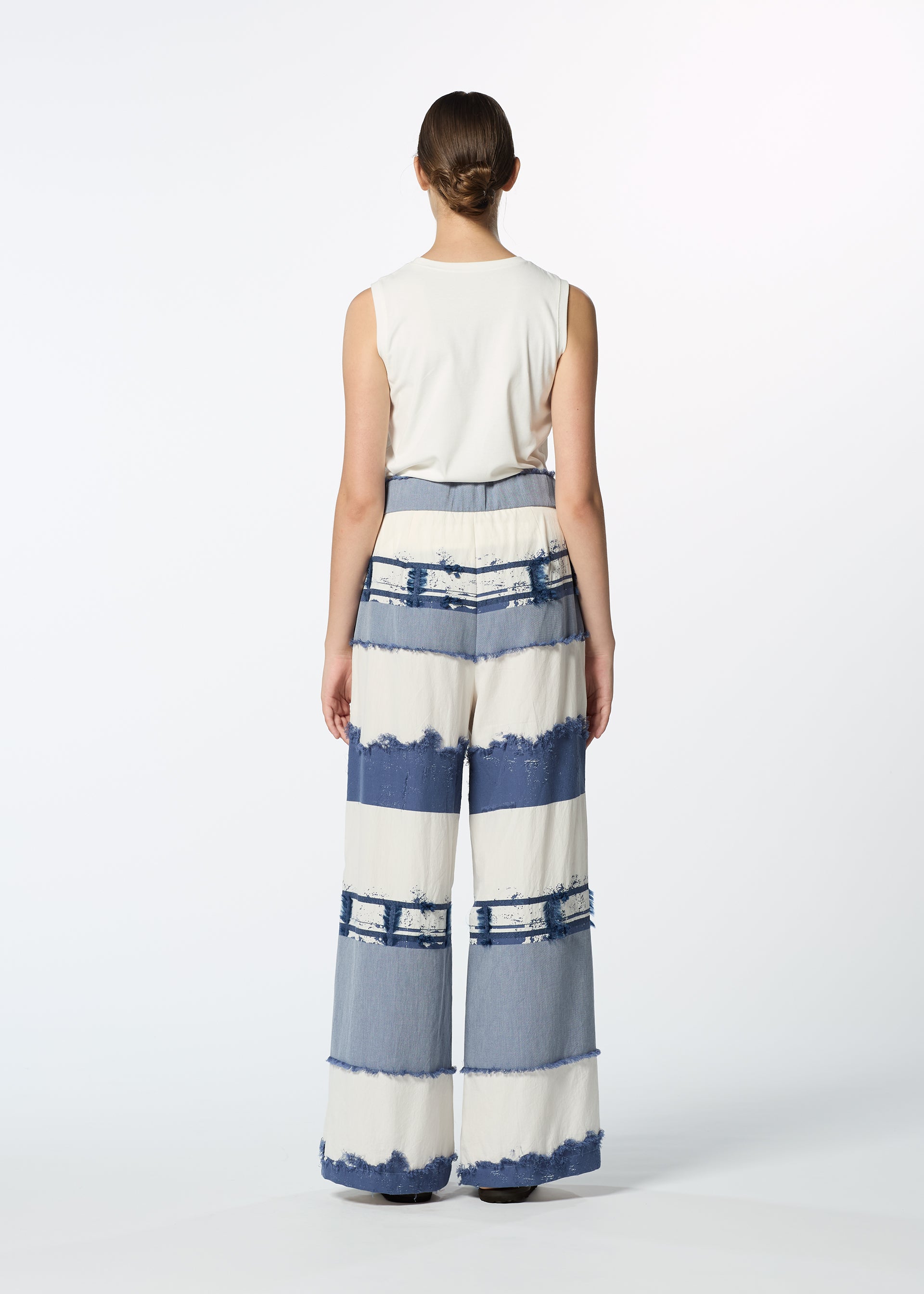 Zoomed view A person stands facing away, wearing HaaT's KASANEGAKE sleeveless white top and jacquard woven wide-leg pants with blue and white stripes, frayed edges, and varied texture, set against a plain white background.
