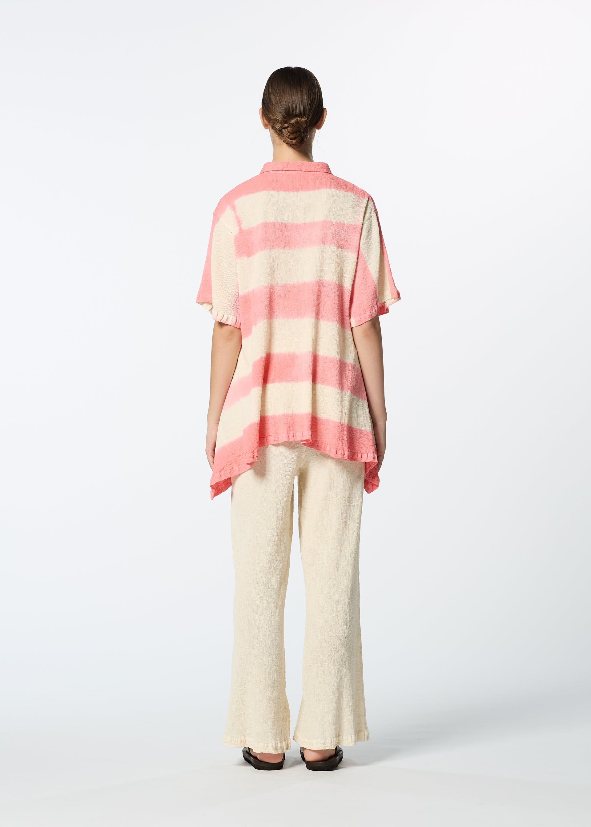 Zoomed view A person faces away against a white background, wearing a loose, short-sleeved pink and white striped shirt, cream HaaT KYO CHIJIMI ITAJIME pants made from ultra-fine cotton yarn on a unique loom, and black shoes.