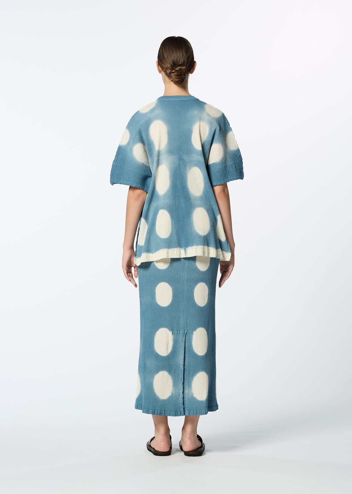 A woman, seen from behind, wears HaaT's KYO CHIJIMI ITAJIME set: a blue top with large white polka dots and a handcrafted skirt, styled with black sandals against a plain white background. view 9 of 9