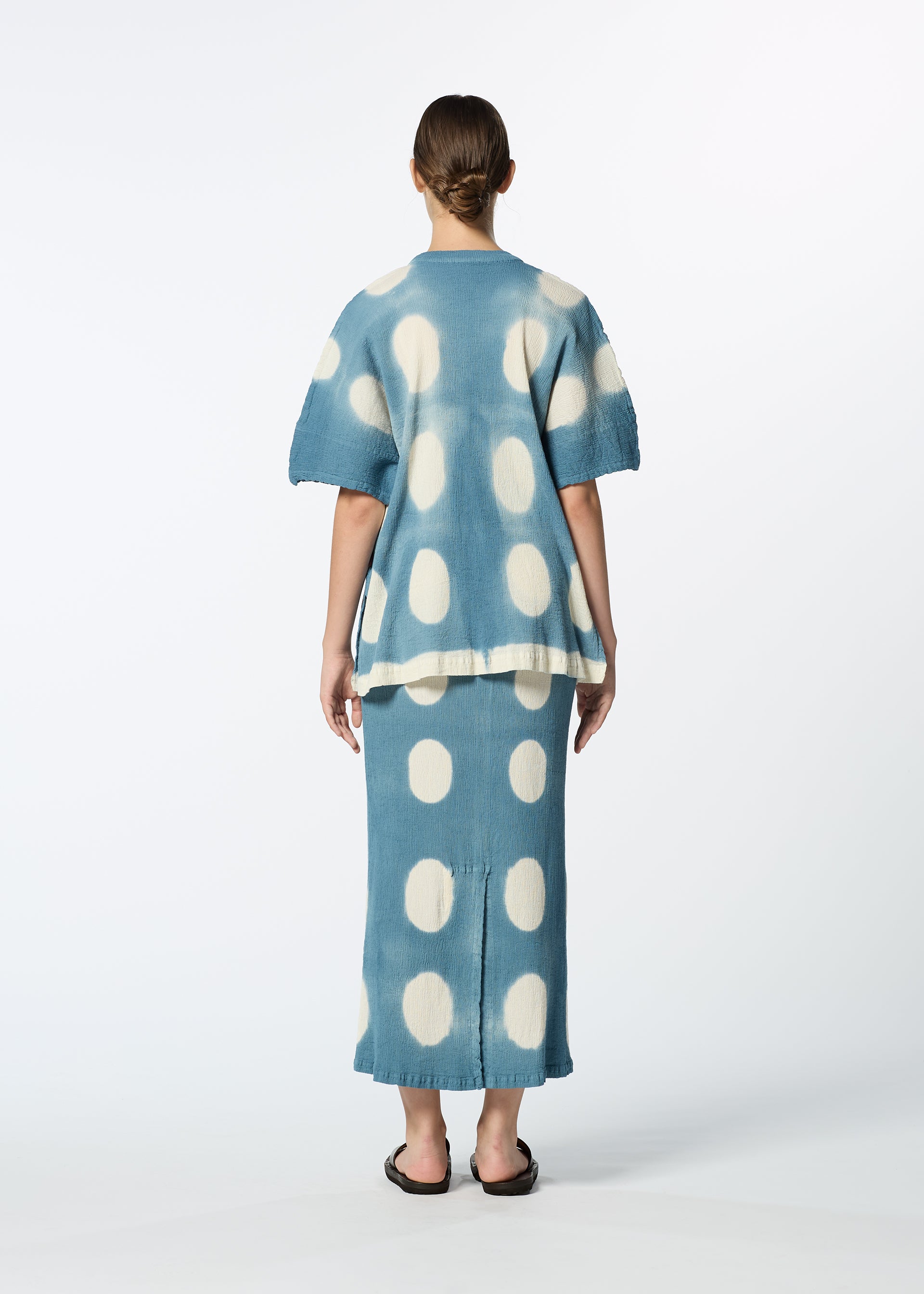Zoomed view A woman, seen from behind, wears HaaT's KYO CHIJIMI ITAJIME set: a blue top with large white polka dots and a handcrafted skirt, styled with black sandals against a plain white background.