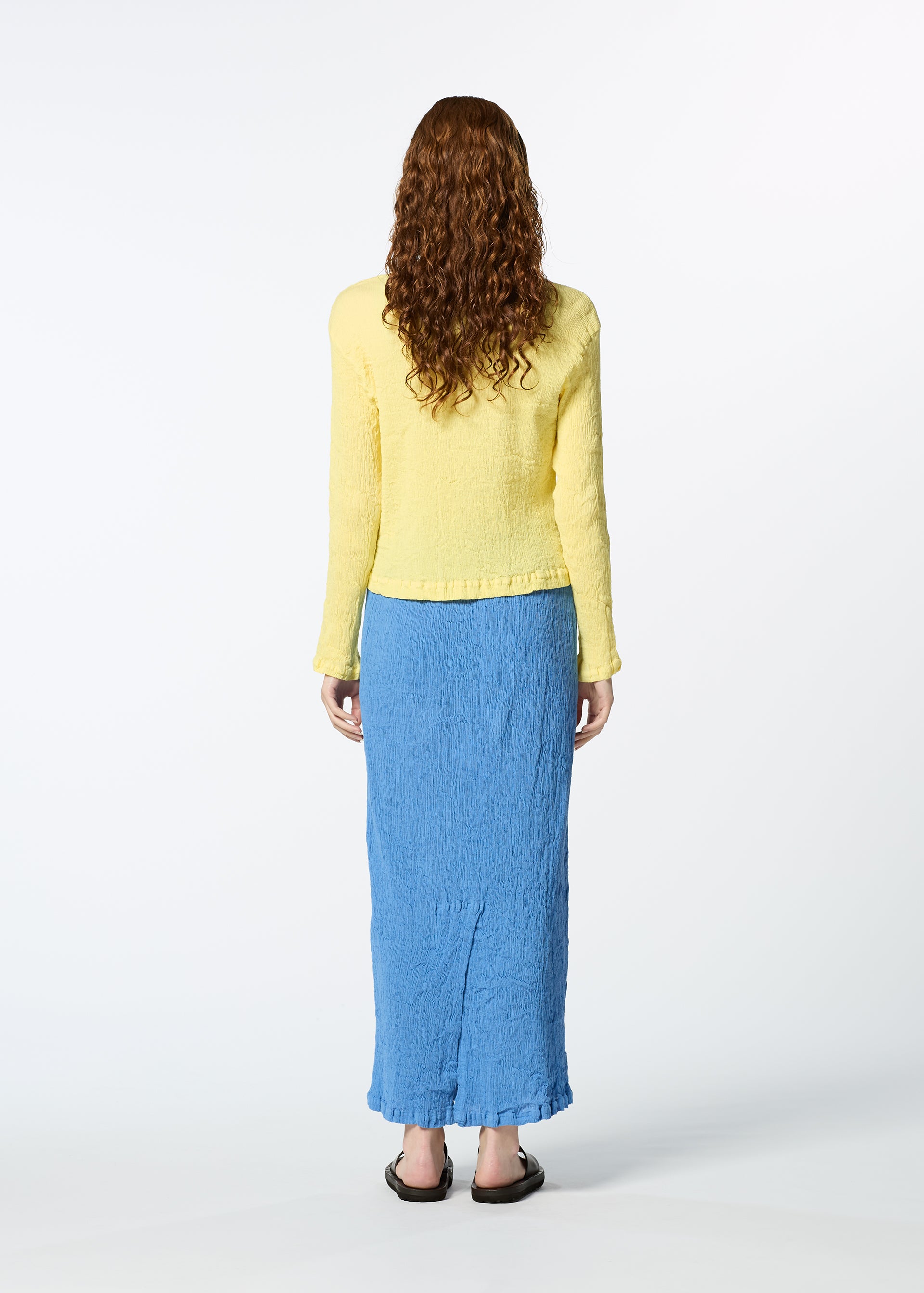Zoomed view A person with long curly hair stands facing away, wearing a yellow long-sleeve top, black sandals, and the HaaT KYO CHIJIMI MAY blue skirt made from ultra-fine cotton yarn, against a plain white background.