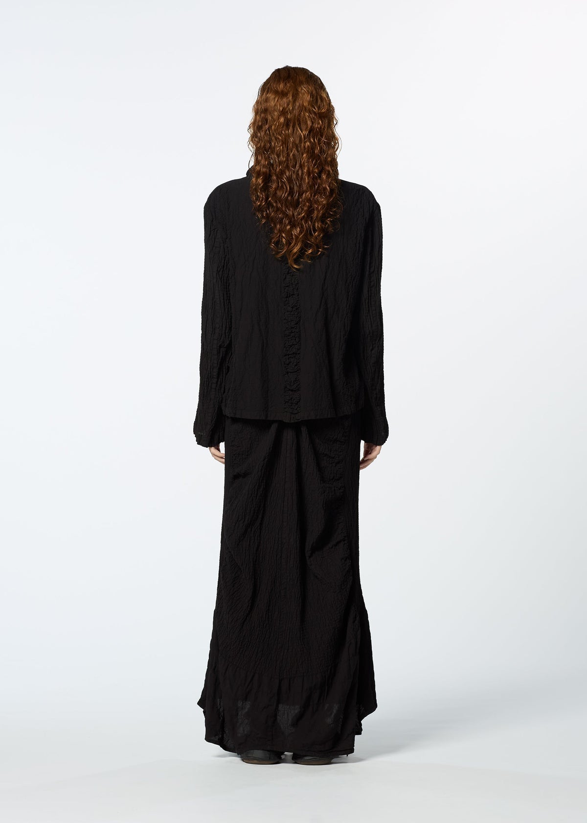 A person with long, curly hair stands facing away, wearing HaaT's TENMOKU COTTON loose black jacket and a garment-dyed textured skirt against a plain white background. view 7 of 7