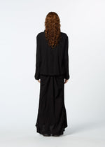 A person with long, curly hair stands facing away, wearing HaaT's TENMOKU COTTON loose black jacket and a garment-dyed textured skirt against a plain white background.