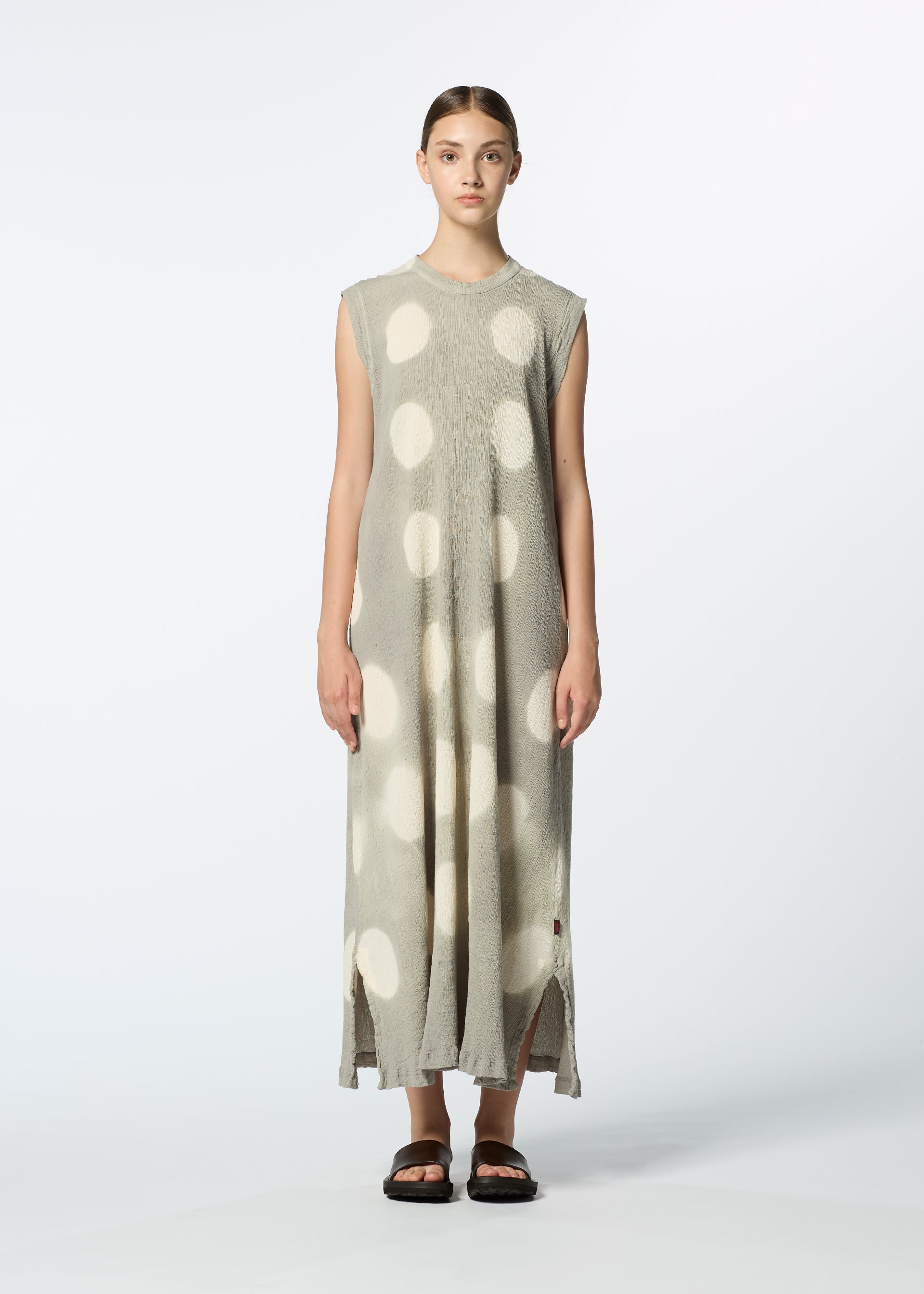 Zoomed view A woman faces forward wearing a loose, sleeveless gray HaaT KYO CHIJIMI ITAJIME dress with large white polka dots, slit hems, and black sandals against a plain white background.