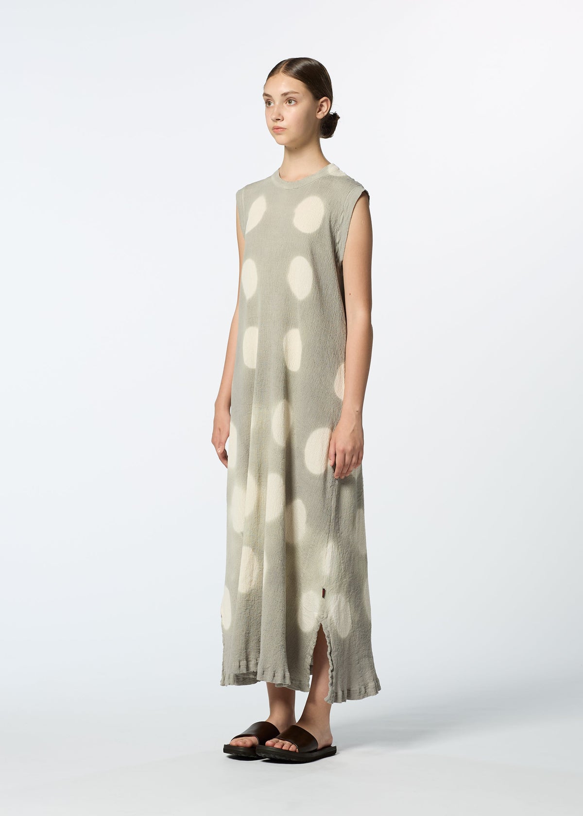 A person stands in a studio wearing HaaT's KYO CHIJIMI ITAJIME, a sleeveless light gray handcrafted dress with large white polka dots, side slits at the hem, paired with black sandals against a plain white background. view 7 of 8