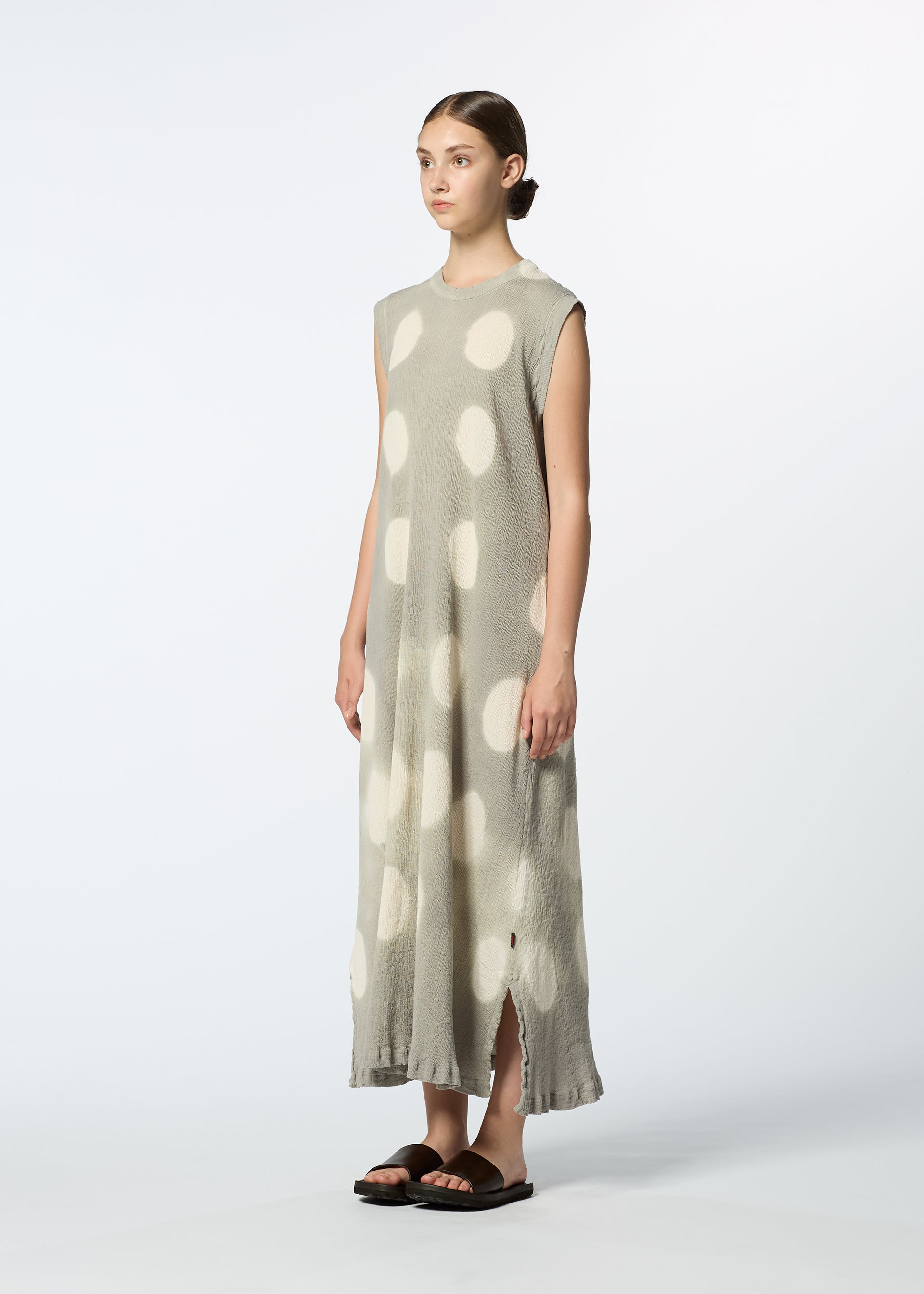 Zoomed view A person stands in a studio wearing HaaT's KYO CHIJIMI ITAJIME, a sleeveless light gray handcrafted dress with large white polka dots, side slits at the hem, paired with black sandals against a plain white background.