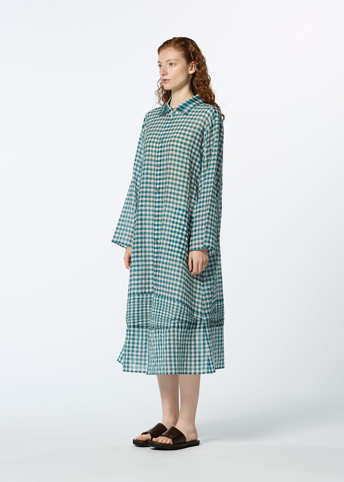 A woman with curly hair models HaaT's 2 SIDE PINTUCKING long, loose-fitting blue and green gingham dress with long sleeves and black sandals, standing against a plain white background. view 7 of 8