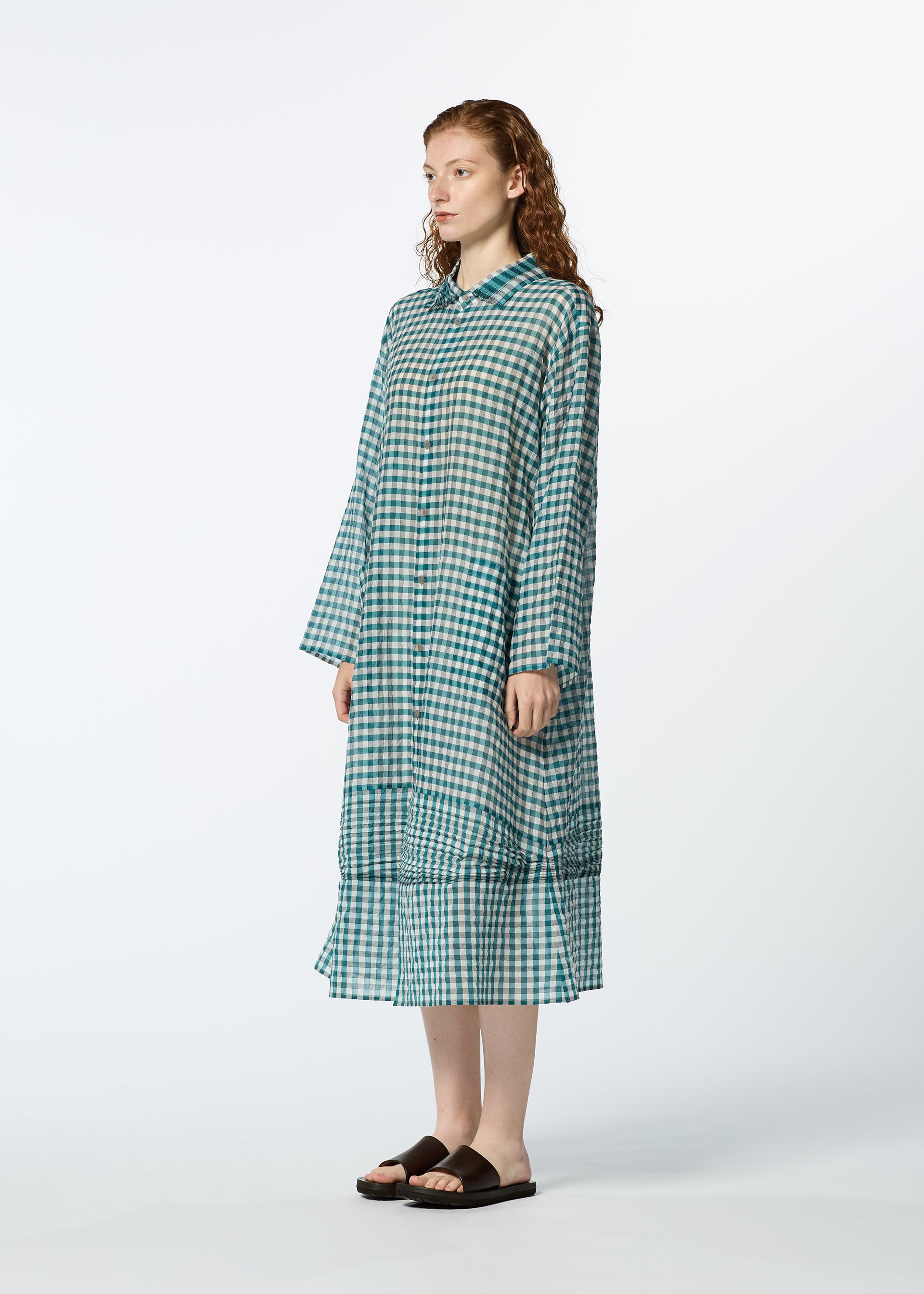 Zoomed view A woman with curly hair models HaaT's 2 SIDE PINTUCKING long, loose-fitting blue and green gingham dress with long sleeves and black sandals, standing against a plain white background.