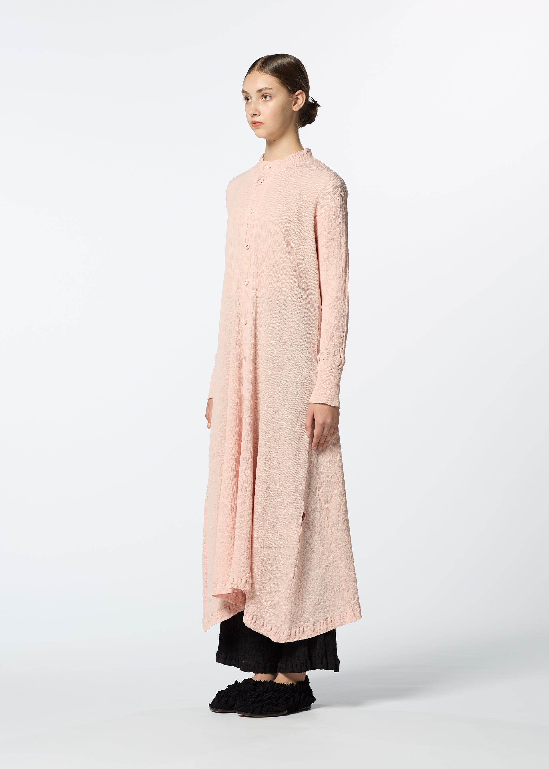 Zoomed view A woman with her hair pulled back stands against a light background, wearing HaaT’s KYO CHIJIMI FEBRUARY long-sleeved dress over a black skirt and black shoes, facing slightly to the left.