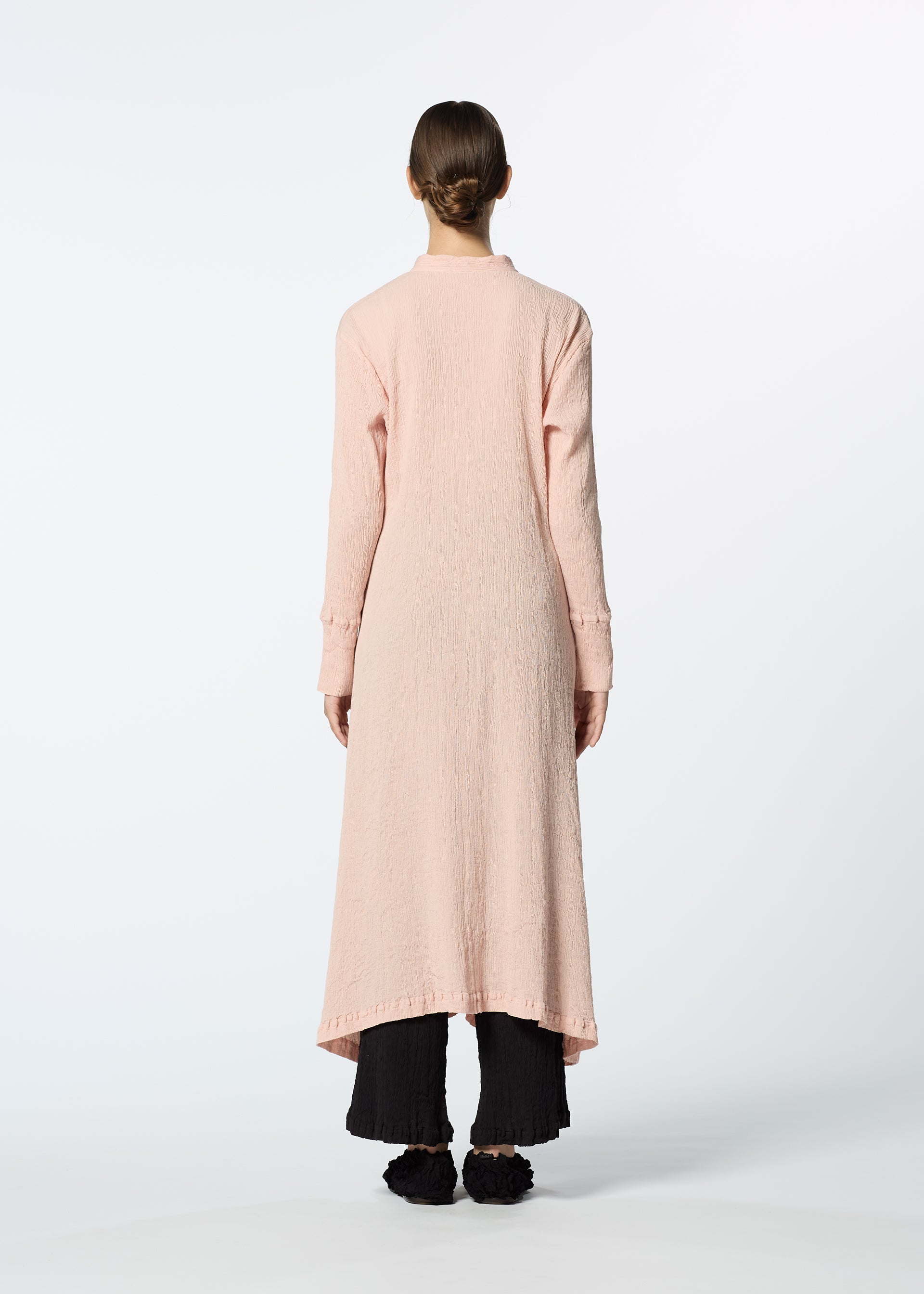 Zoomed view A person stands facing away, wearing the HaaT KYO CHIJIMI FEBRUARY long-sleeved dress made from ultra-fine cotton yarn, layered over wide-leg black pants and black shoes, set against a white background.