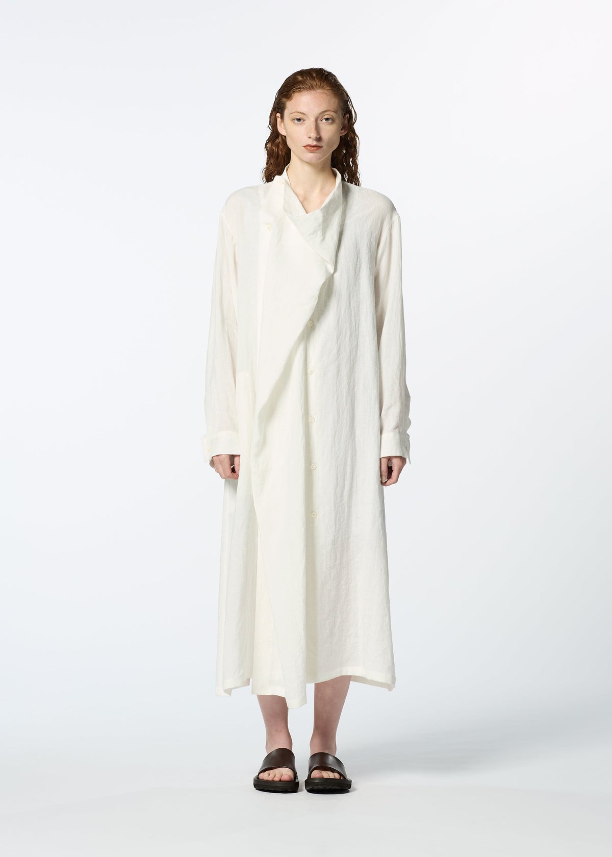 A person with light skin and wavy brown hair stands against a plain white background, wearing HaaT’s JIKI—a long, loose white French linen dress with an asymmetrical draped panel and long sleeves—paired with black slide sandals. view 6 of 9