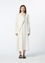 A person with light skin and wavy brown hair stands against a plain white background, wearing HaaT’s JIKI—a long, loose white French linen dress with an asymmetrical draped panel and long sleeves—paired with black slide sandals.