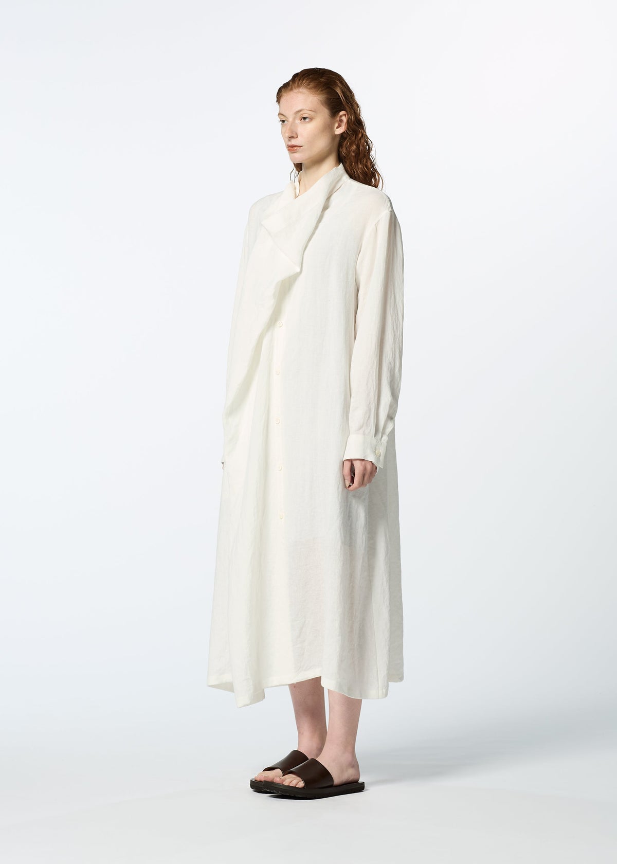 A person with long, reddish hair stands against a plain background, wearing HaaT's JIKI French linen dress with long sleeves for an elegant silhouette, paired with dark flat sandals. view 7 of 9