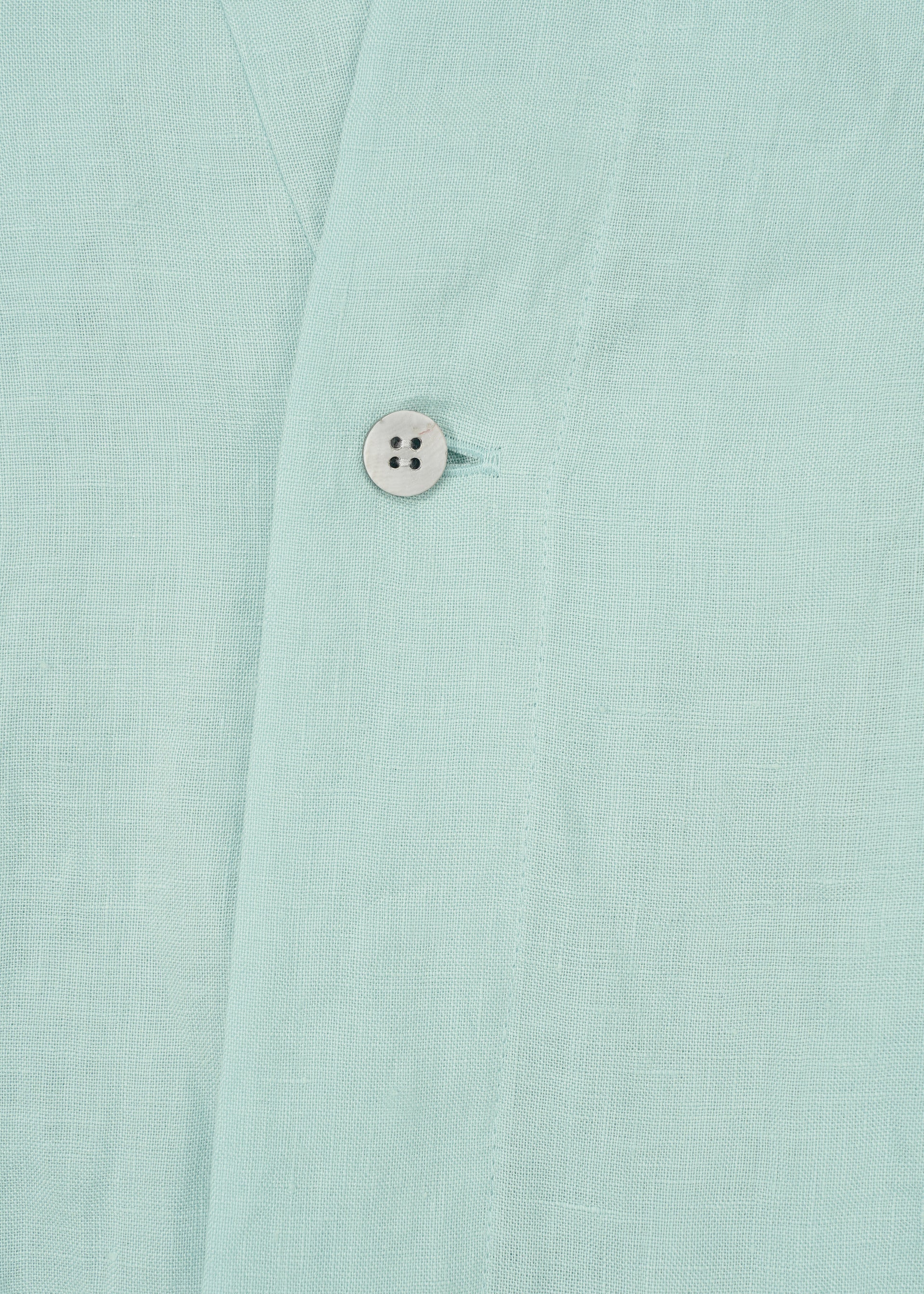 Zoomed view Close-up of JIKI by HaaT: light blue, breathable dress fabric featuring a visible seam and a single white button. The textured, linen-like material suggests an elegant silhouette and refined craftsmanship.