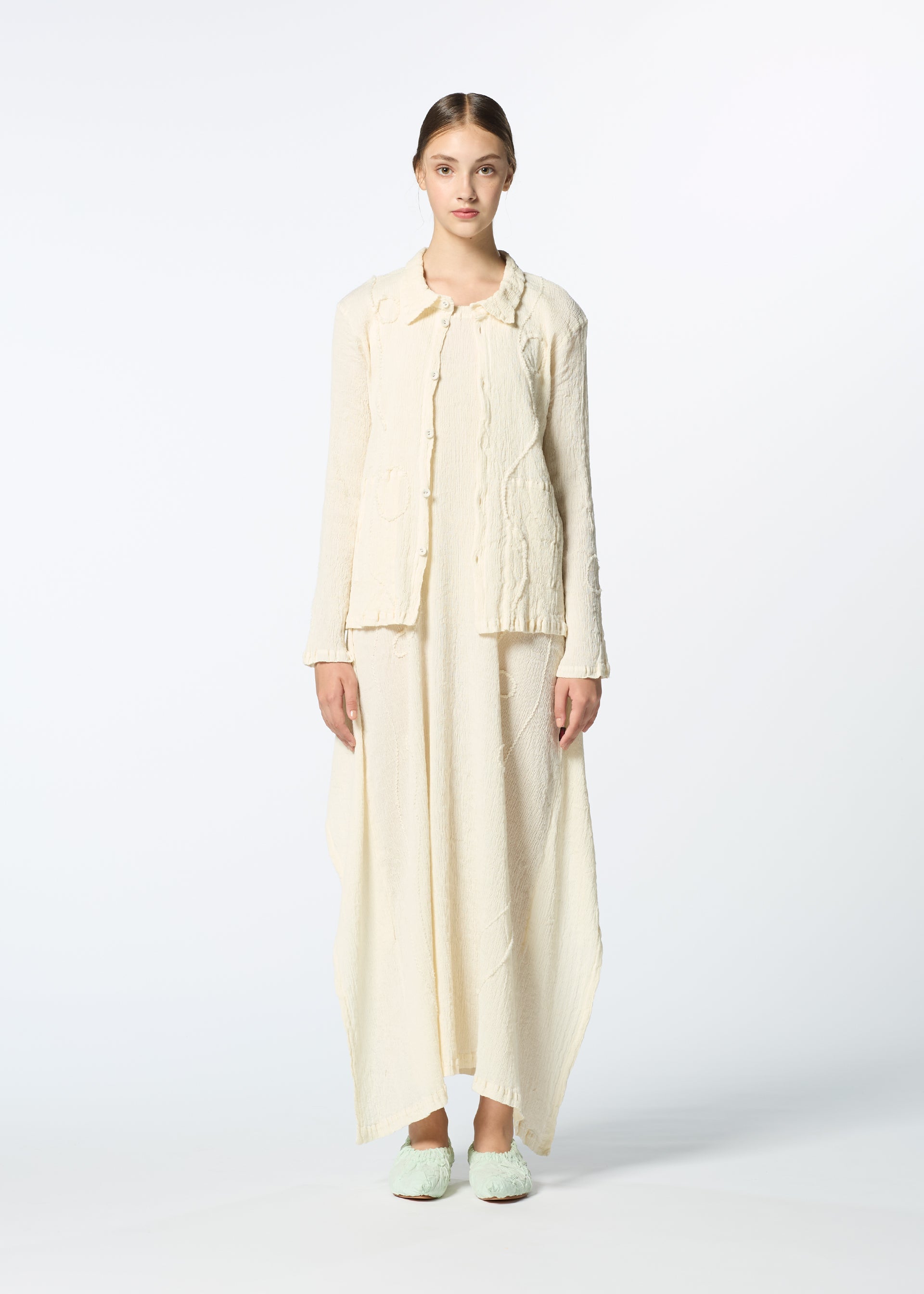 Zoomed view A woman stands facing forward in HaaT’s KYO CHIJIMI EMB set: an off-white, textured long jacket and matching wide ankle-length pants with subtle curved embroidery. Her hair is pulled back, and she wears soft slippers against a plain white background.