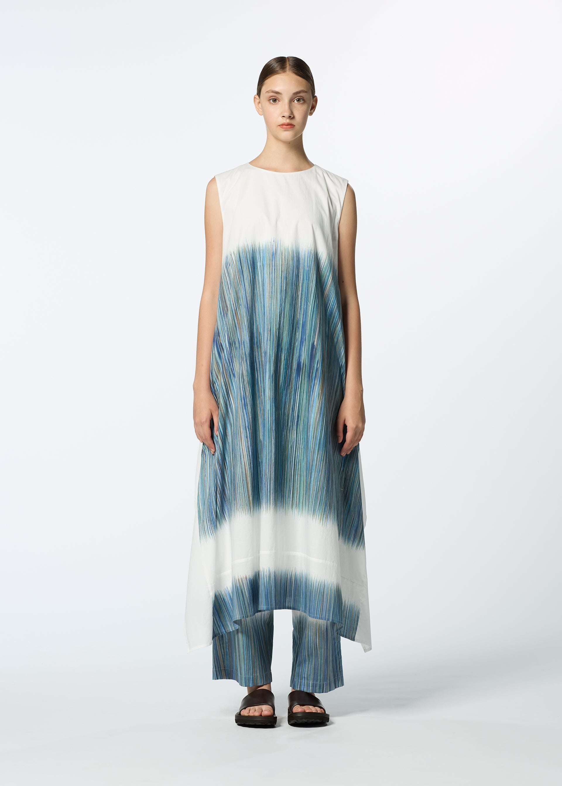 Zoomed view A person stands facing forward wearing HaaT's SAZANAMI—a sleeveless, flowing hand-painted dress with blue, green, and teal vertical streaks—over matching pants and black sandals against a plain light background.