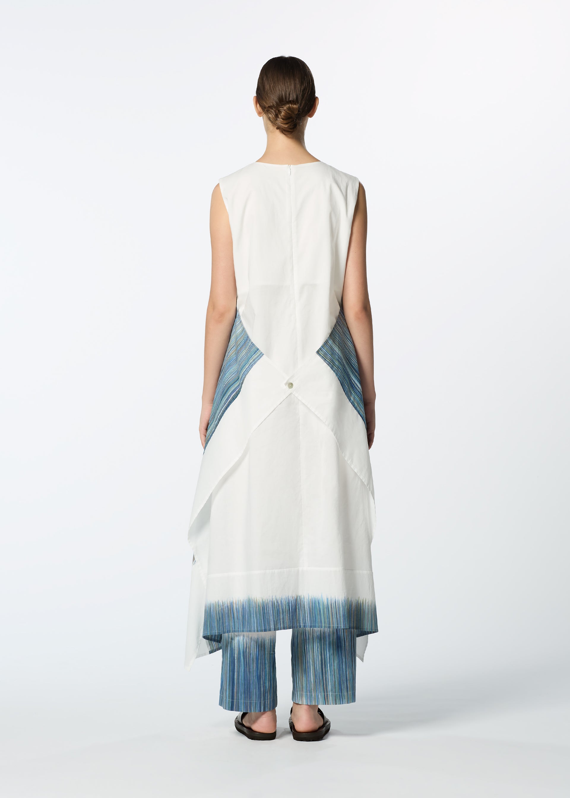 Zoomed view A person stands facing away, wearing HaaT’s SAZANAMI: a sleeveless white organic cotton dress with layered, asymmetrical panels and blue vertical-striped accents over matching striped pants and black sandals against a plain white backdrop.