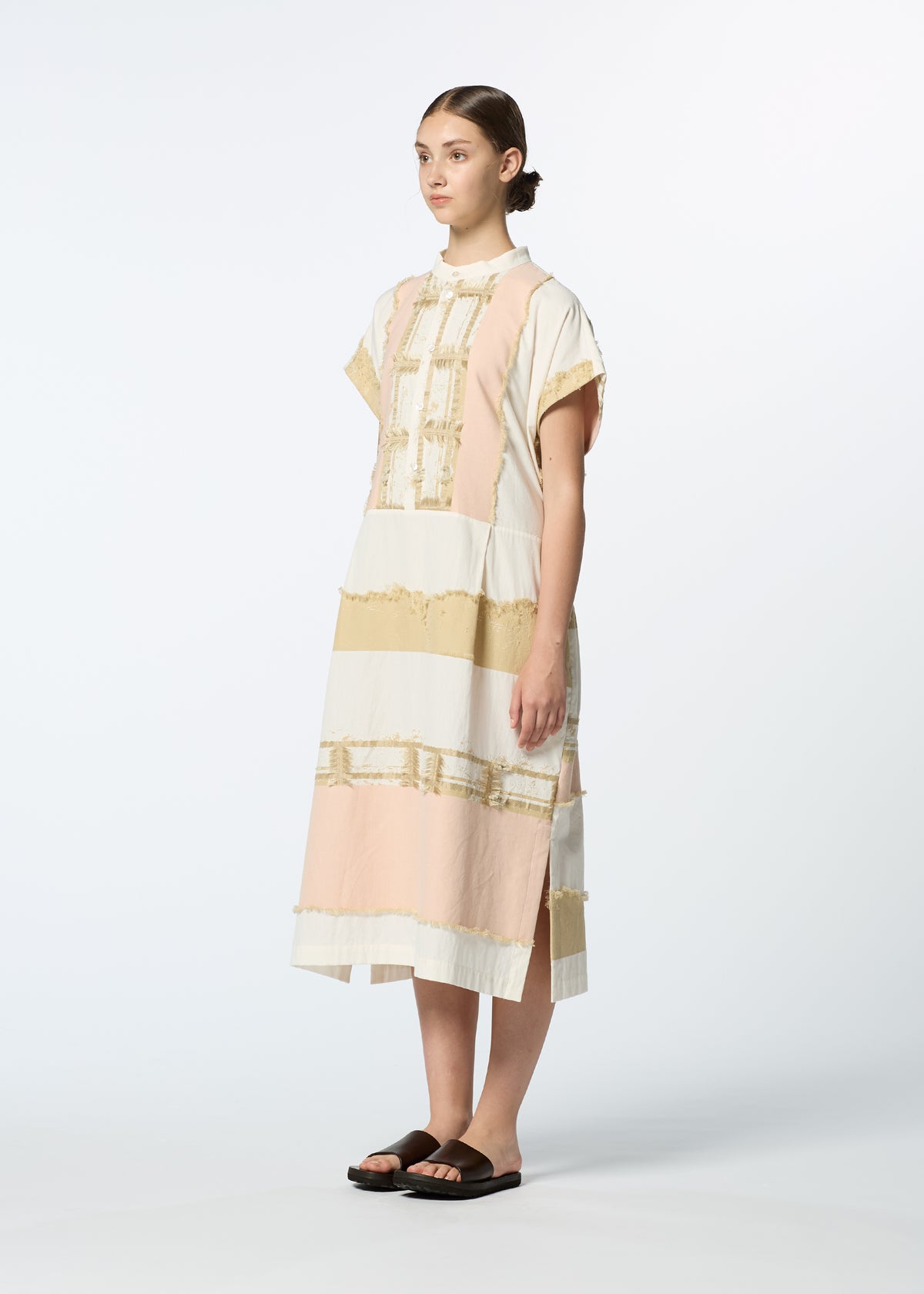 A woman in the HaaT KASANEGAKE jacquard dress with pastel pink, white, and yellow bands and frayed edges stands sideways, her hair slicked back and wearing black sandals against a plain white background. view 7 of 8