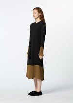 A person with long, wavy red hair wears HaaT’s KYO CHIJIMI STREAM dress, a loose, long-sleeved colored piece with brown cuffs and hem, paired with black slip-on shoes, standing sideways against a plain light background.