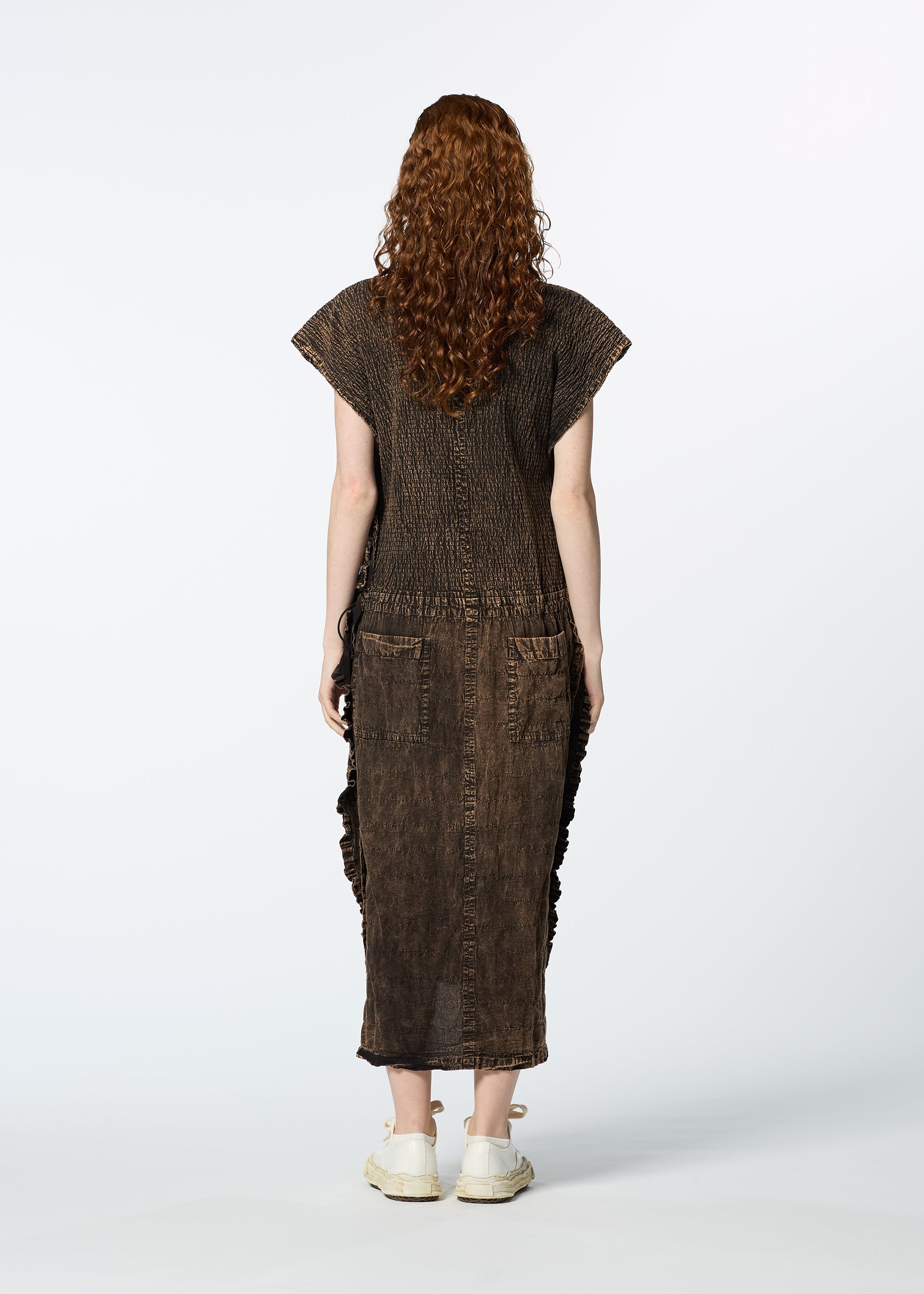 Zoomed view A person with long curly hair, dressed in HaaT's TENMOKU COTTON brown textured garment-dyed dress and white shoes, stands facing away from the camera against a plain white background.