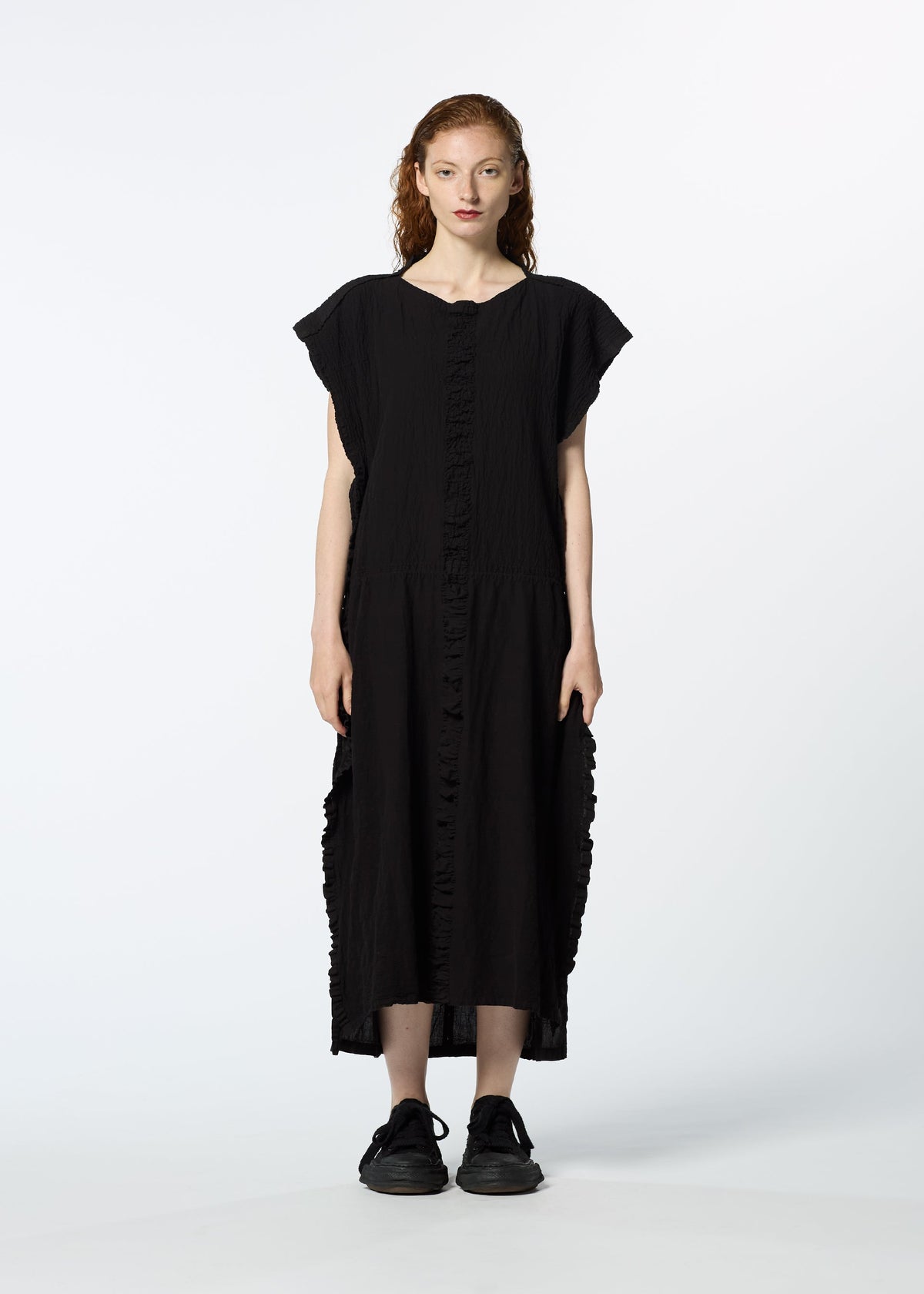 A woman with long, wavy red hair faces forward wearing HaaT’s TENMOKU COTTON, a loose-fit black midi dress with ruffle details and a mottled garment-dye pattern, paired with black sneakers against a plain white background. view 5 of 7