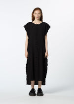 A woman with long, wavy red hair faces forward wearing HaaT’s TENMOKU COTTON, a loose-fit black midi dress with ruffle details and a mottled garment-dye pattern, paired with black sneakers against a plain white background.