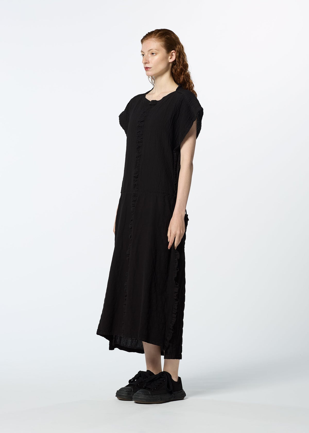 A person with long red hair stands sideways, wearing HaaT's TENMOKU COTTON—a loose-fitting black midi dress with a subtle mottled pattern inspired by Tenmoku glaze—paired with black sneakers, against a plain white background. view 6 of 7